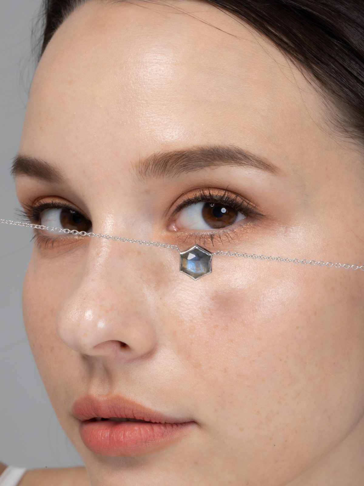 Close-up of a woman's face with a silver chain and labradorite gemstone on a grey background