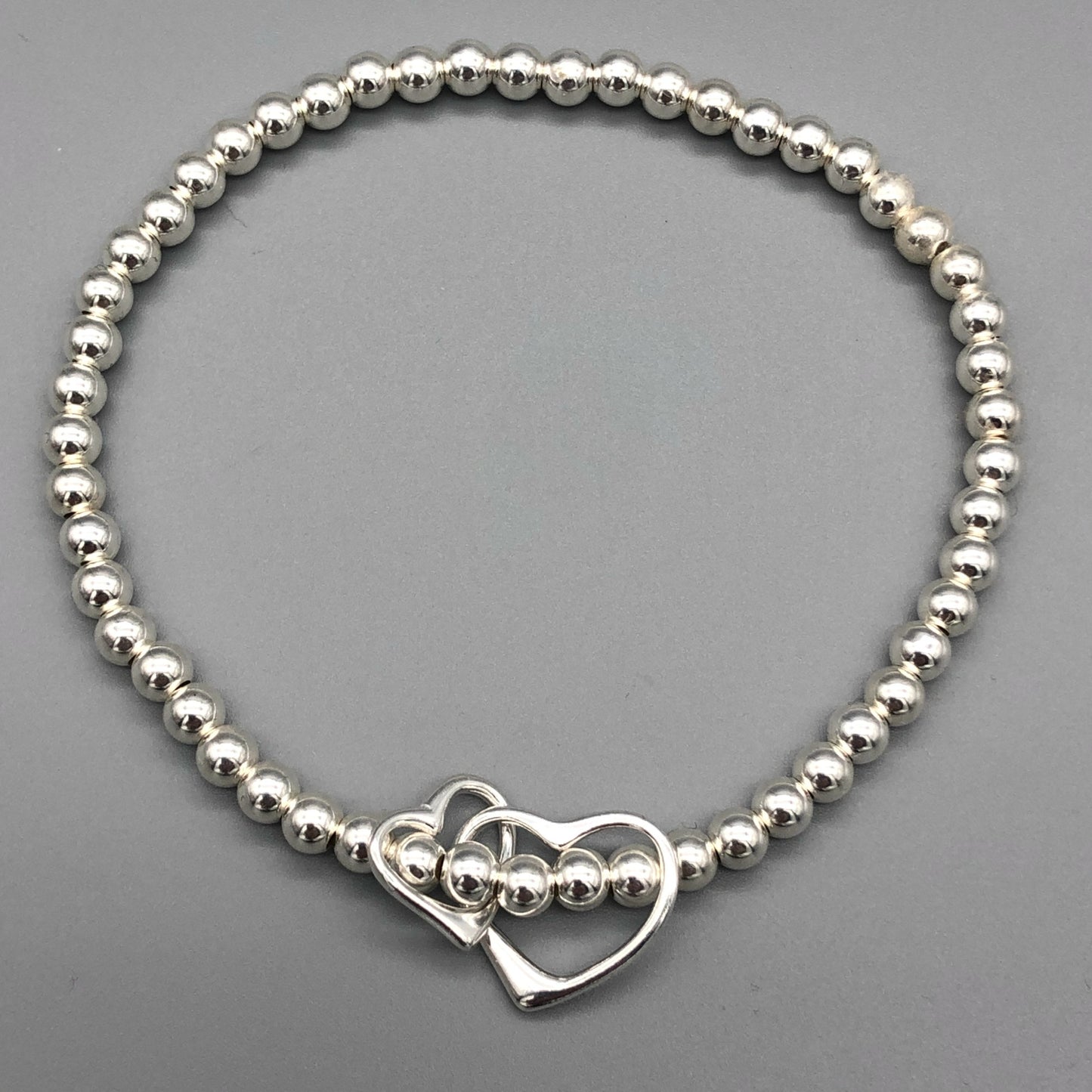 A sterling silver bracelet featuring a double interlinked open heart charm as the central element, with small silver beads on stretch elastic by My Silver Wish