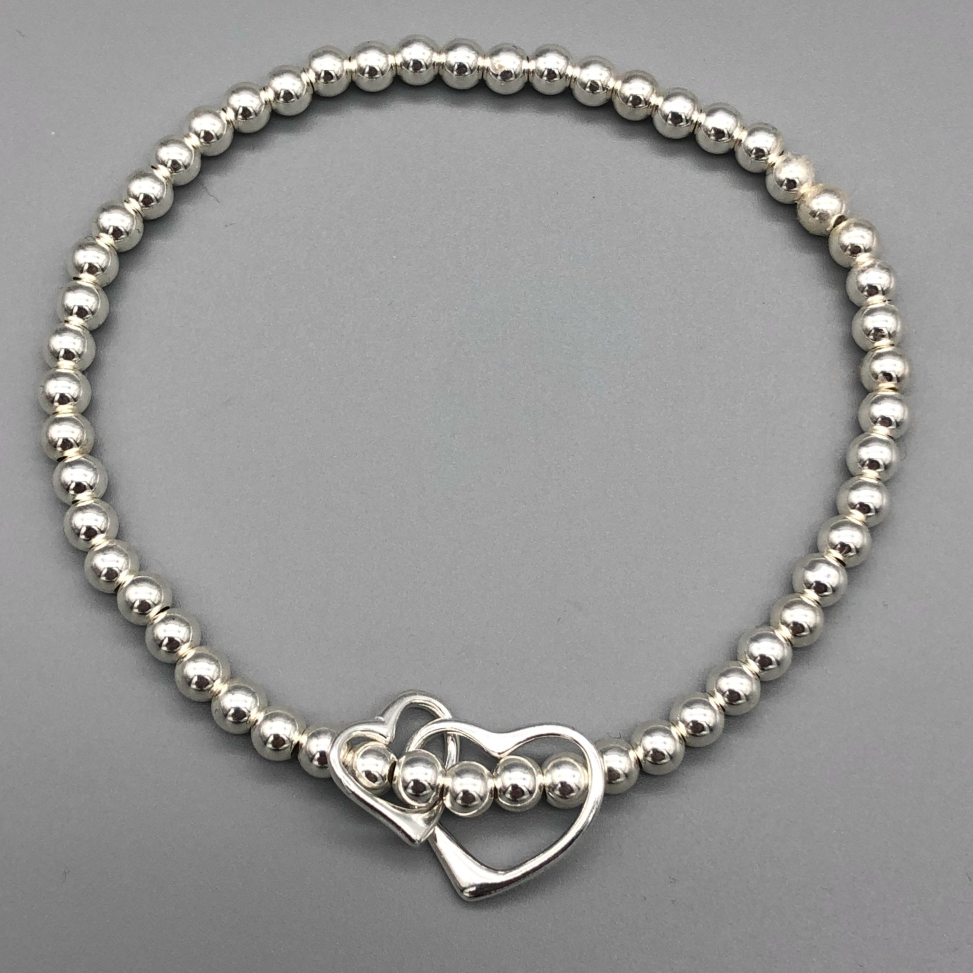 A sterling silver bracelet featuring a double interlinked open heart charm as the central element, with small silver beads on stretch elastic by My Silver Wish